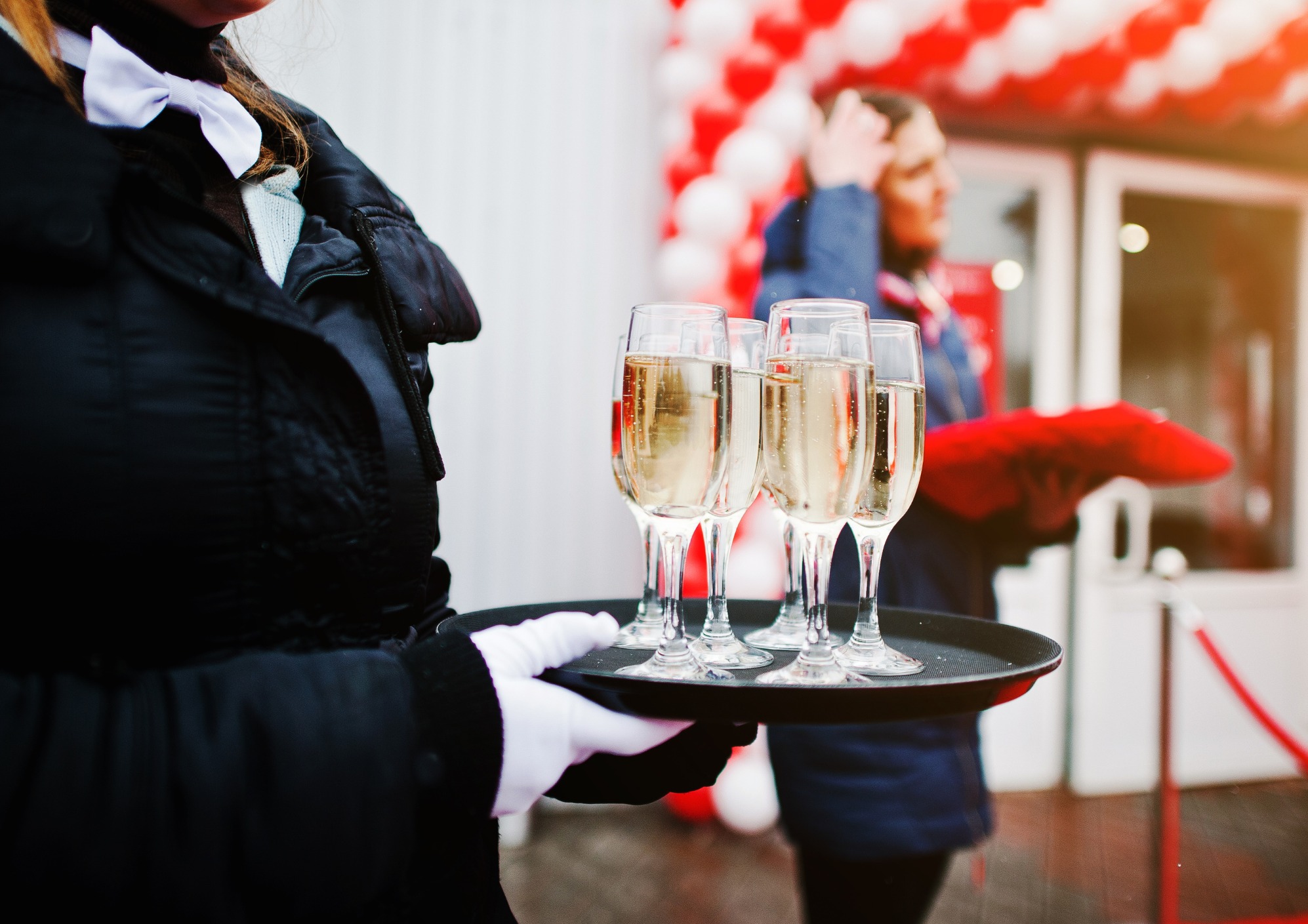 Kelner w białych rękawiczkach i muszce trzyma tacę z kieliszkami szampana na tle wejścia udekorowanego czerwono-białymi balonami.