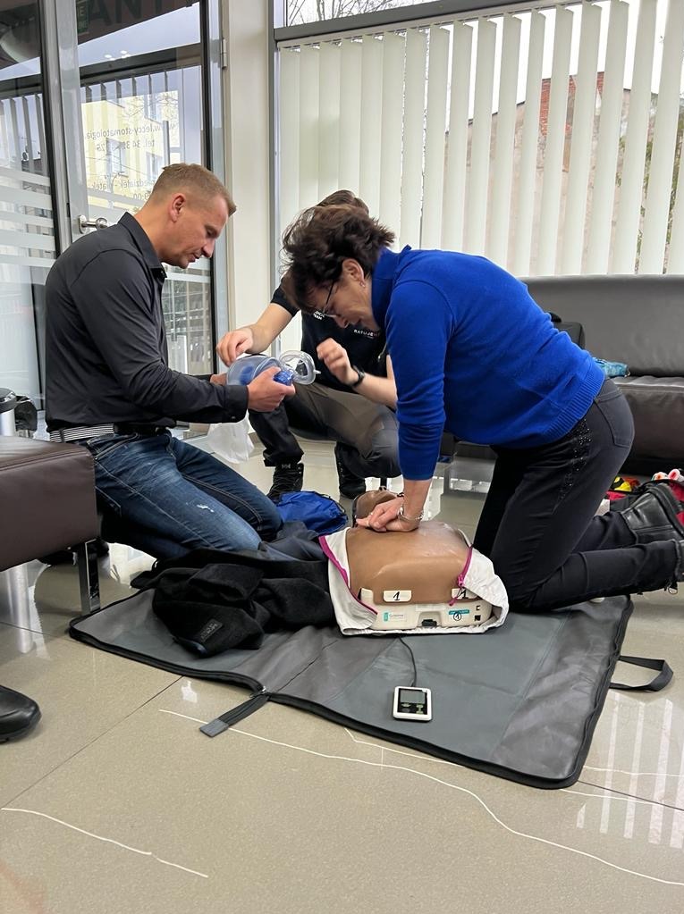 Osoby ćwiczą resuscytację krążeniowo-oddechową na manekinie z użyciem maski resuscytacyjnej i defibrylatora w pomieszczeniu z żaluzjami.