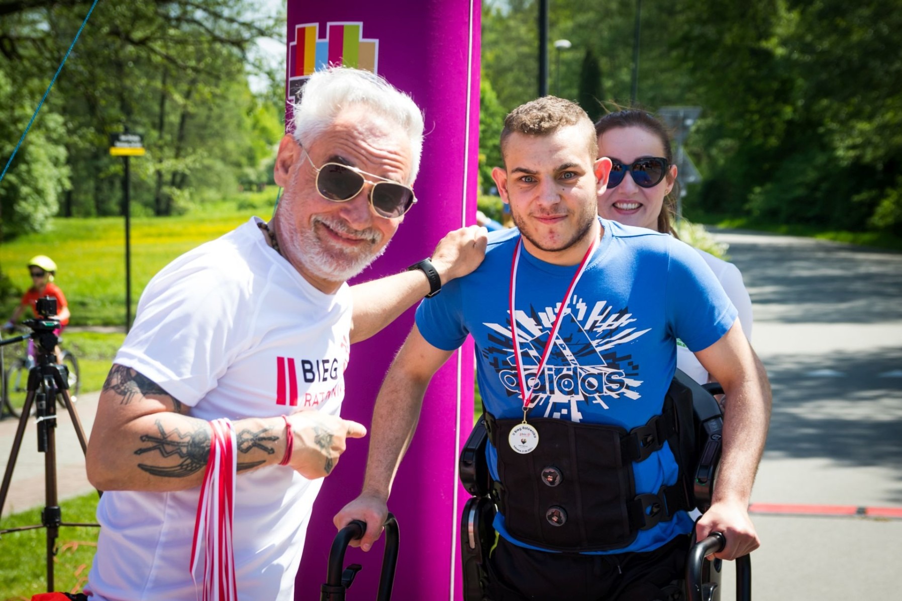 Uśmiechnięty mężczyzna w okularach słonecznych i koszulce z napisem 'Bieg Ratunkowy' wspiera młodego mężczyznę na wózku inwalidzkim, który ma na sobie medal po biegu, w tle kobieta również...