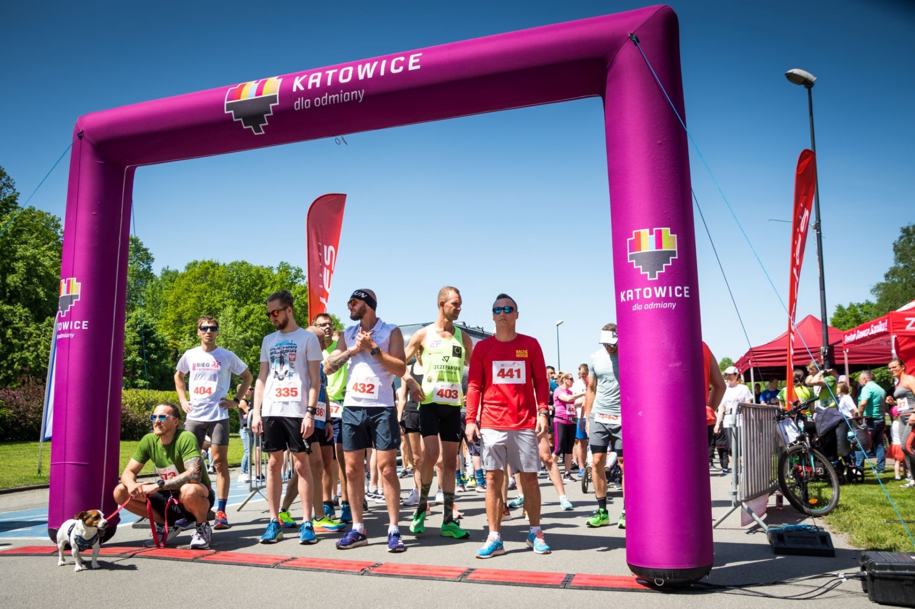 Grupa biegaczy ustawiona na linii startu pod fioletowym dmuchanym łukiem z napisem Katowice dla odmiany, w tle drzewa i błękitne niebo, na pierwszym planie mężczyzna w okularach słonecznych kuca...