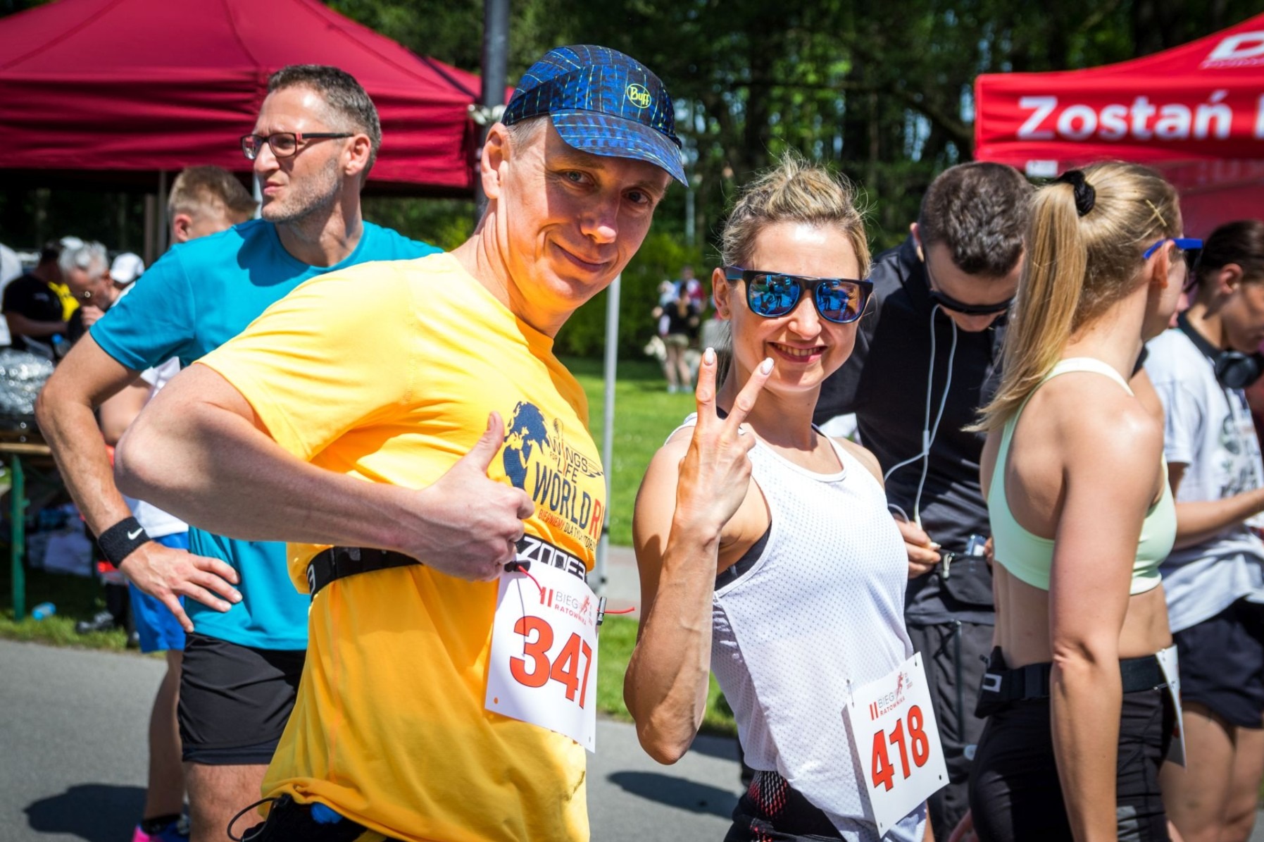 Uczestnicy biegu Wings for Life World Run z numerami startowymi, uśmiechnięci i pokazujący gesty, na tle punktu organizacyjnego.