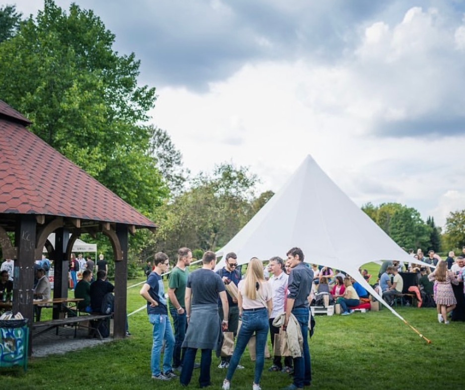 Grupa ludzi w plenerze, stojąca na trawie obok białego namiotu w kształcie gwiazdy i drewnianej altany z czerwonym dachem, w tle drzewa i pochmurne niebo.