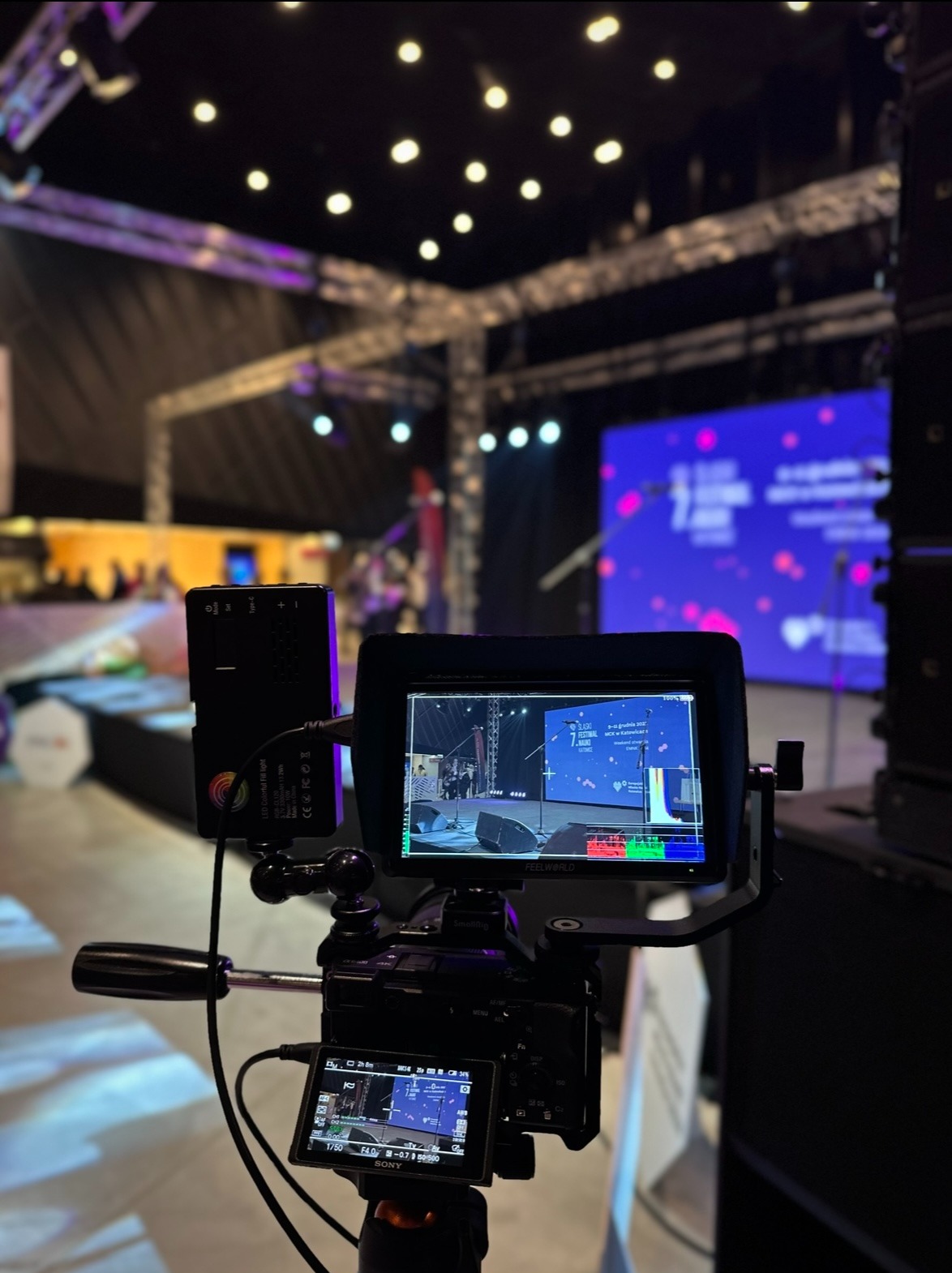 Kamera wideo z podglądem na ekranie, rejestrująca scenę z mikrofonami i ekranem LED podczas wydarzenia; widoczny monitor podglądowy z kolorowymi paskami kalibracyjnymi.