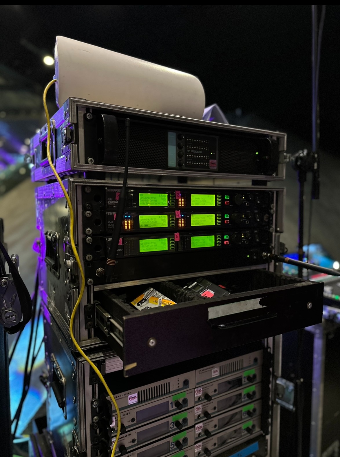 Sprzęt nagłośnieniowy w racku: wzmacniacze, odbiorniki bezprzewodowe mikrofonów Shure i Sennheiser z otwartą szufladą na akcesoria, okablowanie, oświetlenie sceniczne w tle.