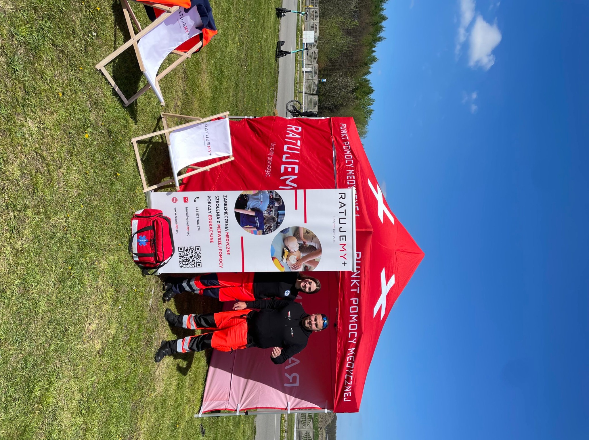 Stanowisko mobilne z czerwonym namiotem i logo 'Ratujemy+', z dwoma ratownikami w strojach oraz torbą medyczną, ustawione na trawie w słoneczny dzień, obok leżaków plażowych.
