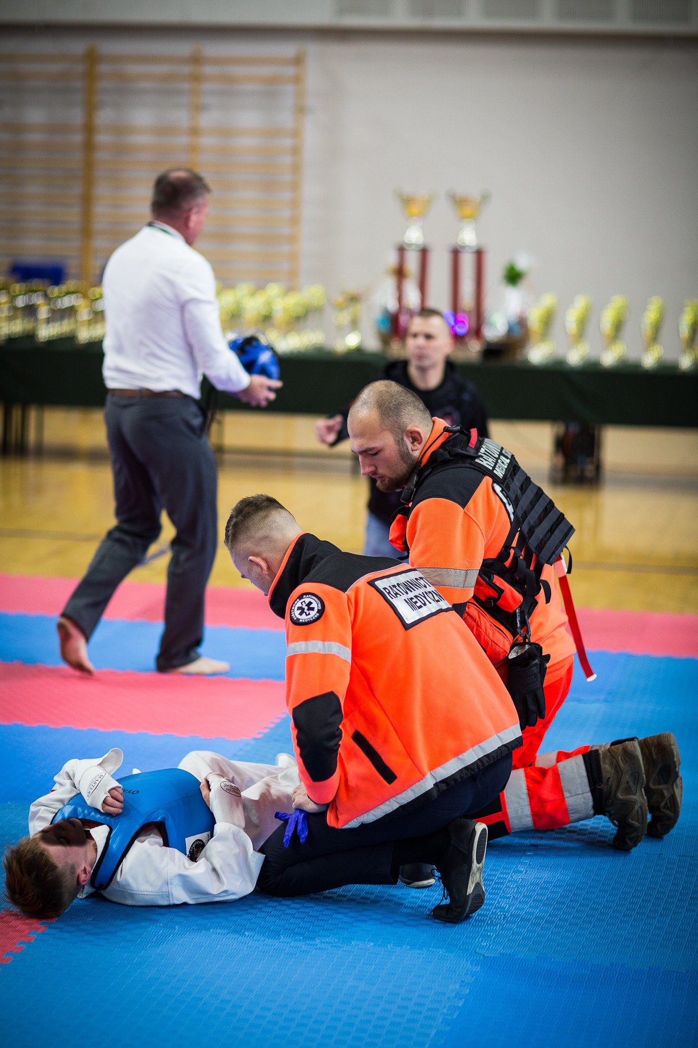 Ratownicy medyczni udzielają pierwszej pomocy zawodnikowi leżącemu na macie, widoczne trofea w tle.