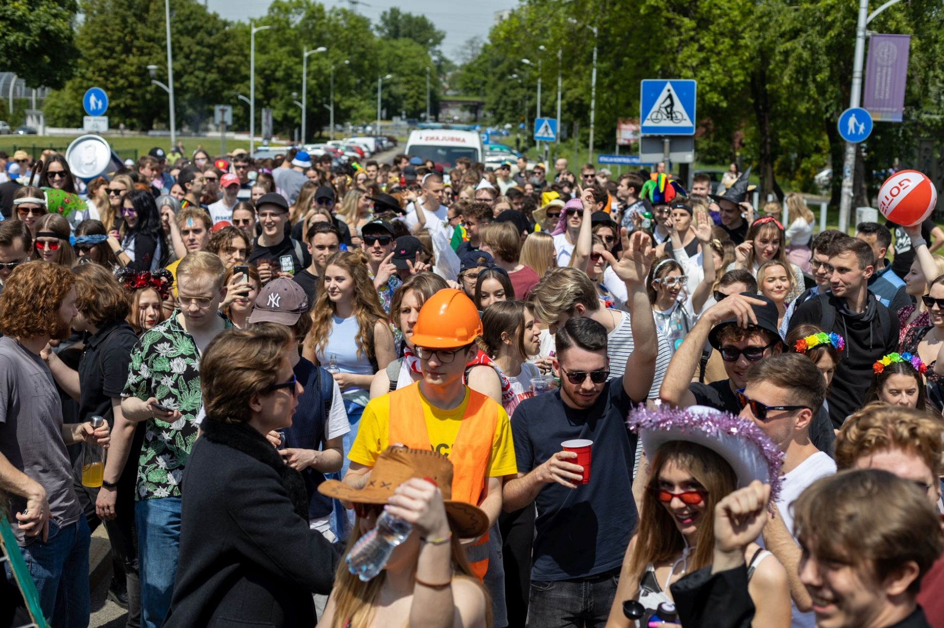 Tłum ludzi na ulicy, niektórzy w kostiumach i przebraniach, w tle drzewa i budynki, widoczne znaki drogowe i ambulans.
