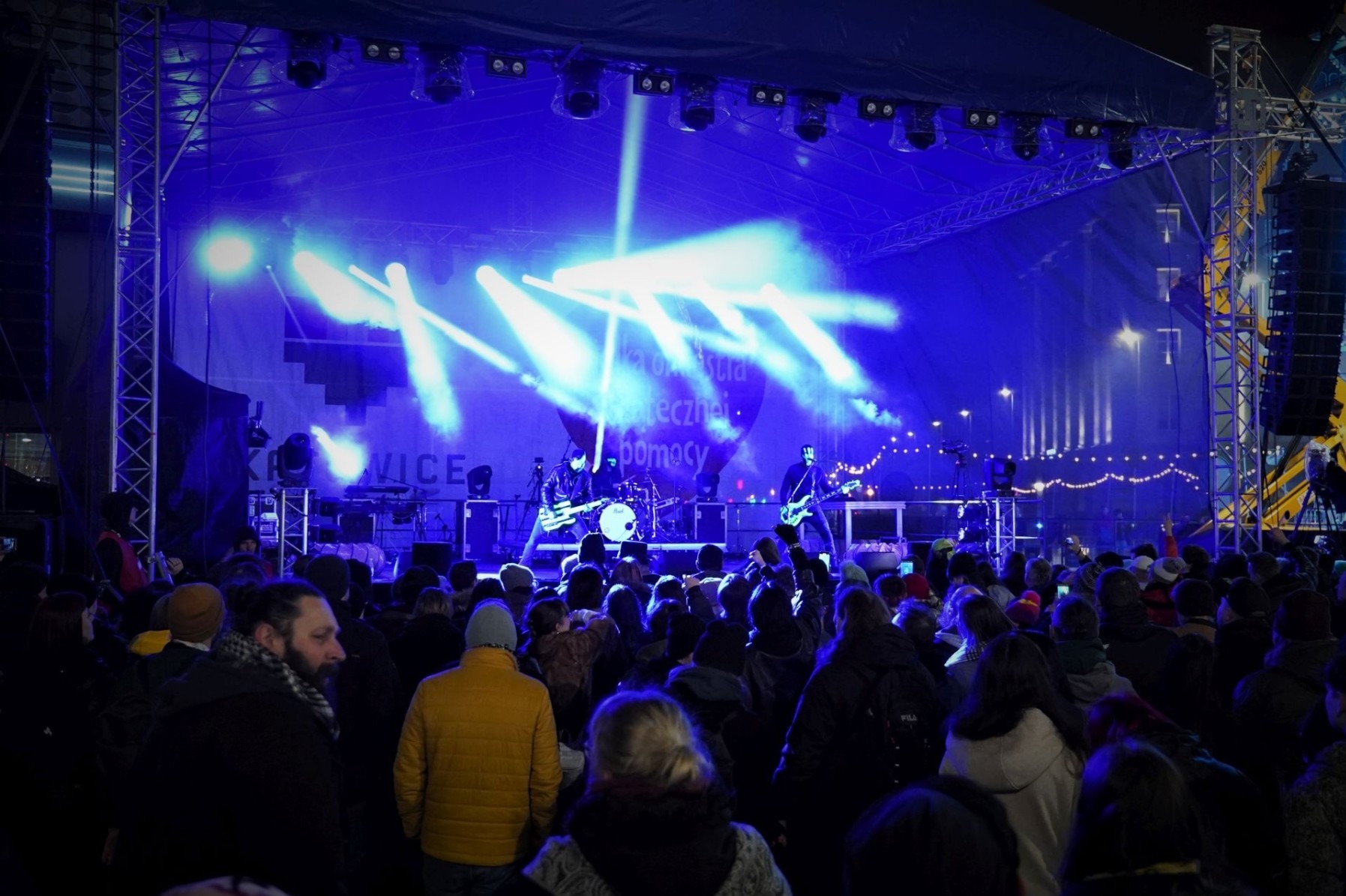 Koncert plenerowy z zespołem na scenie oświetlonej niebieskimi reflektorami, widoczna publiczność i logo Wielkiej Orkiestry Świątecznej Pomocy na tle sceny.