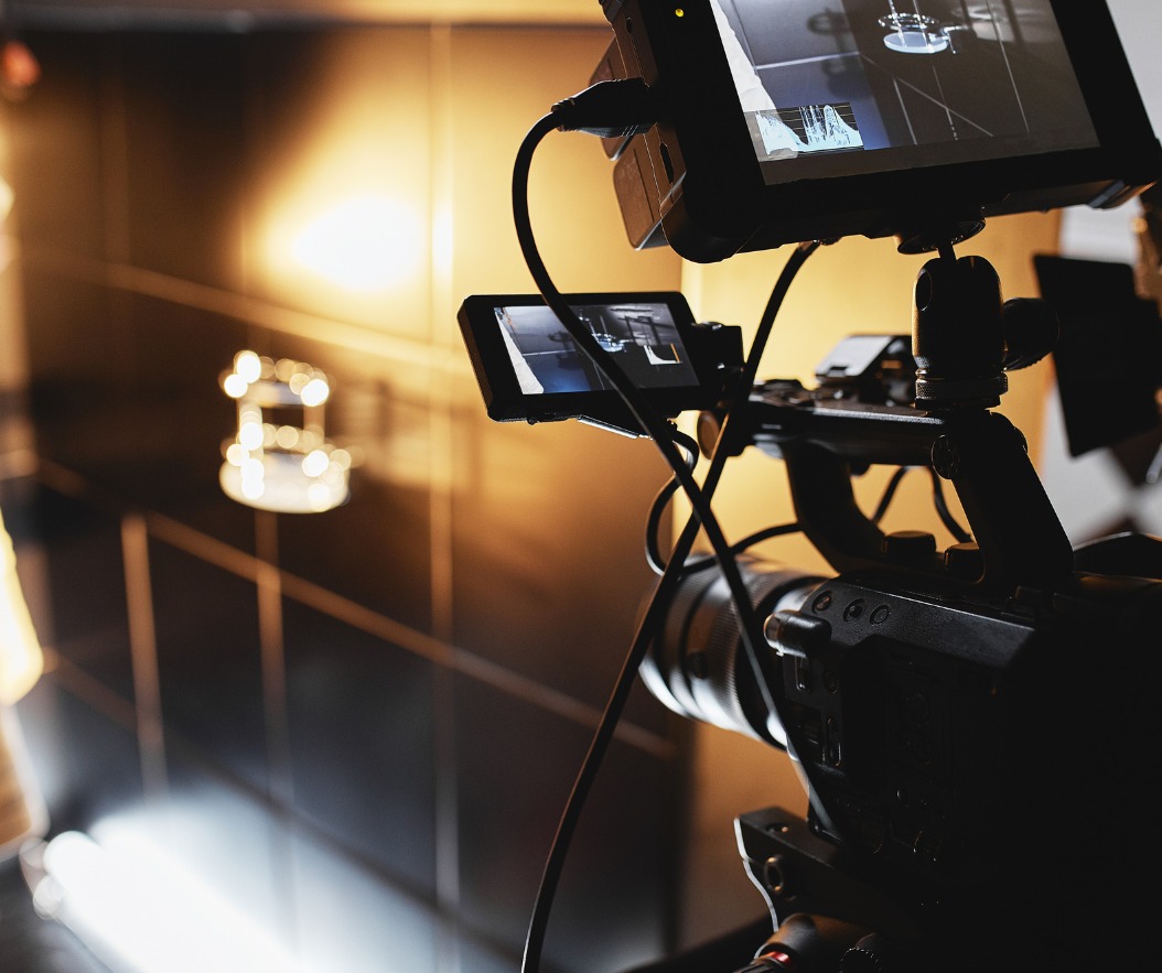 Profesjonalna kamera filmowa z dwoma ekranami podglądu, ustawiona na statywie, skierowana w stronę jasnego, rozmytego tła, prawdopodobnie podczas nagrywania wnętrza lub sceny.