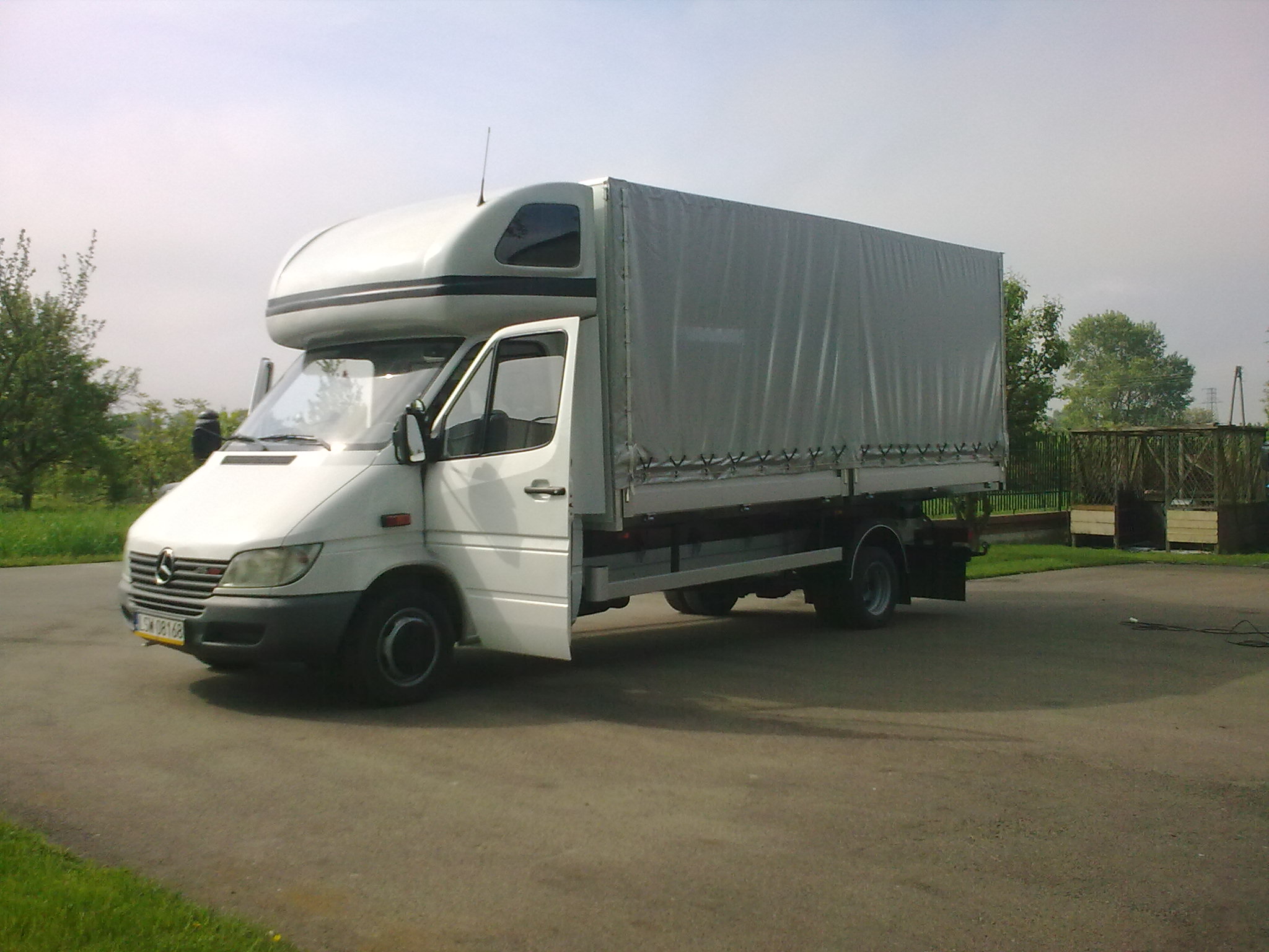 Biały samochód dostawczy z plandeką, zaparkowany w Trawnikach, z otwartymi drzwiami kierowcy, widoczny na tle zieleni i błękitnego nieba.