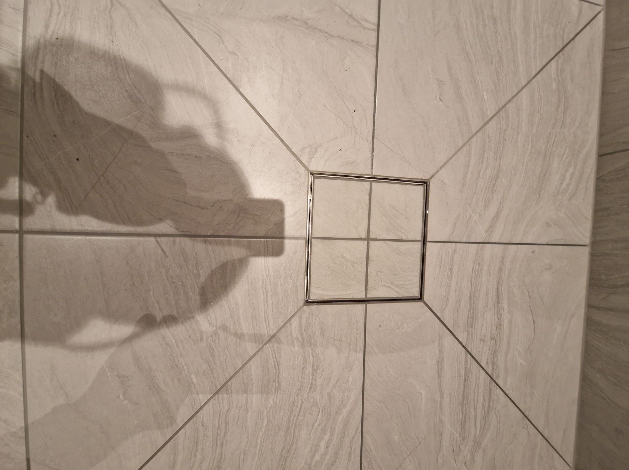 Szare płytki podłogowe ułożone promieniście wokół kwadratowego odpływu liniowego w kabinie prysznicowej, widoczny cień osoby robiącej zdjęcie.