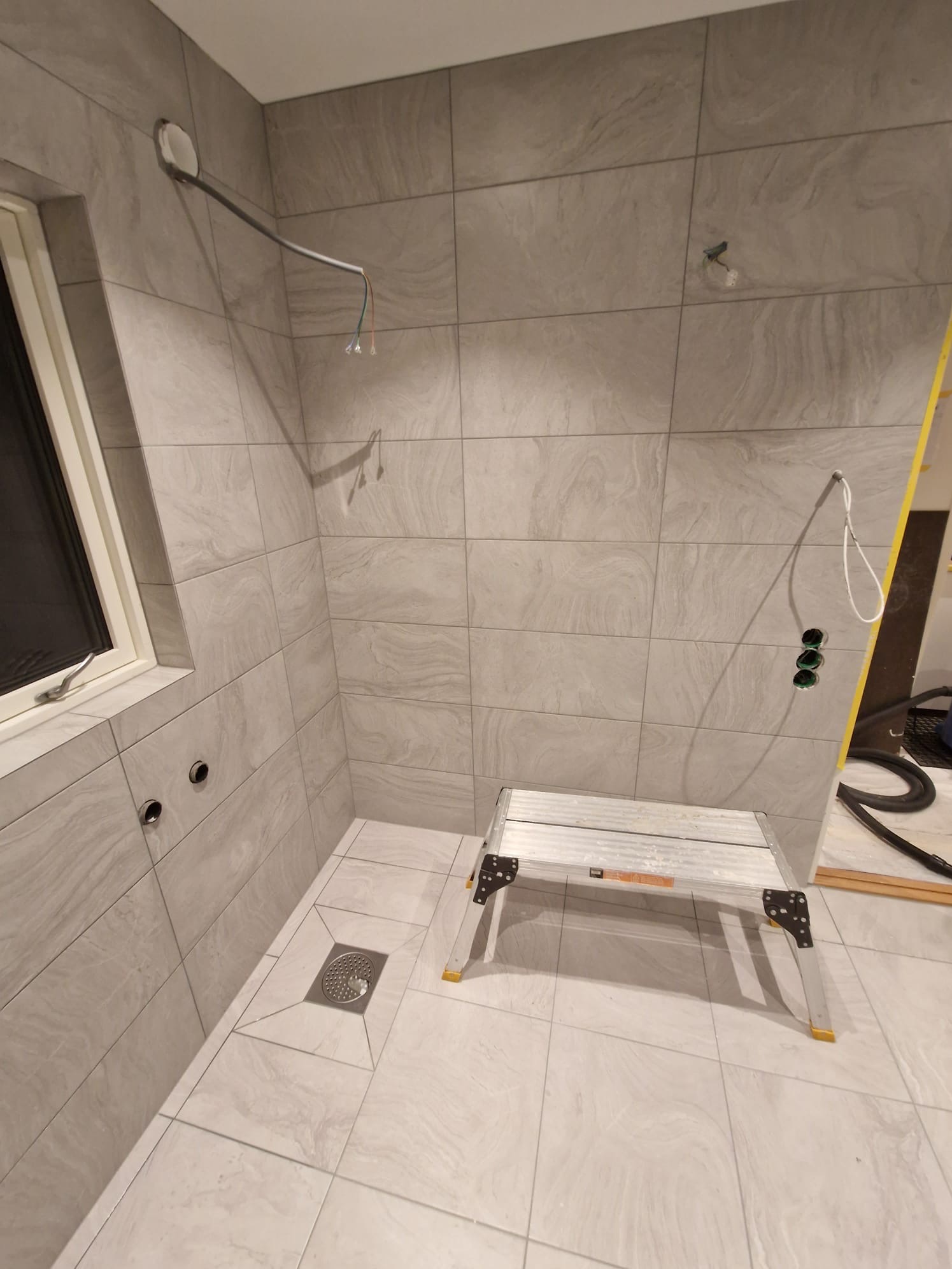 Łazienka w trakcie remontu: szare płytki imitujące kamień na ścianach i podłodze, widoczne instalacje elektryczne i hydrauliczne, drabina aluminiowa, odpływ liniowy w podłodze.