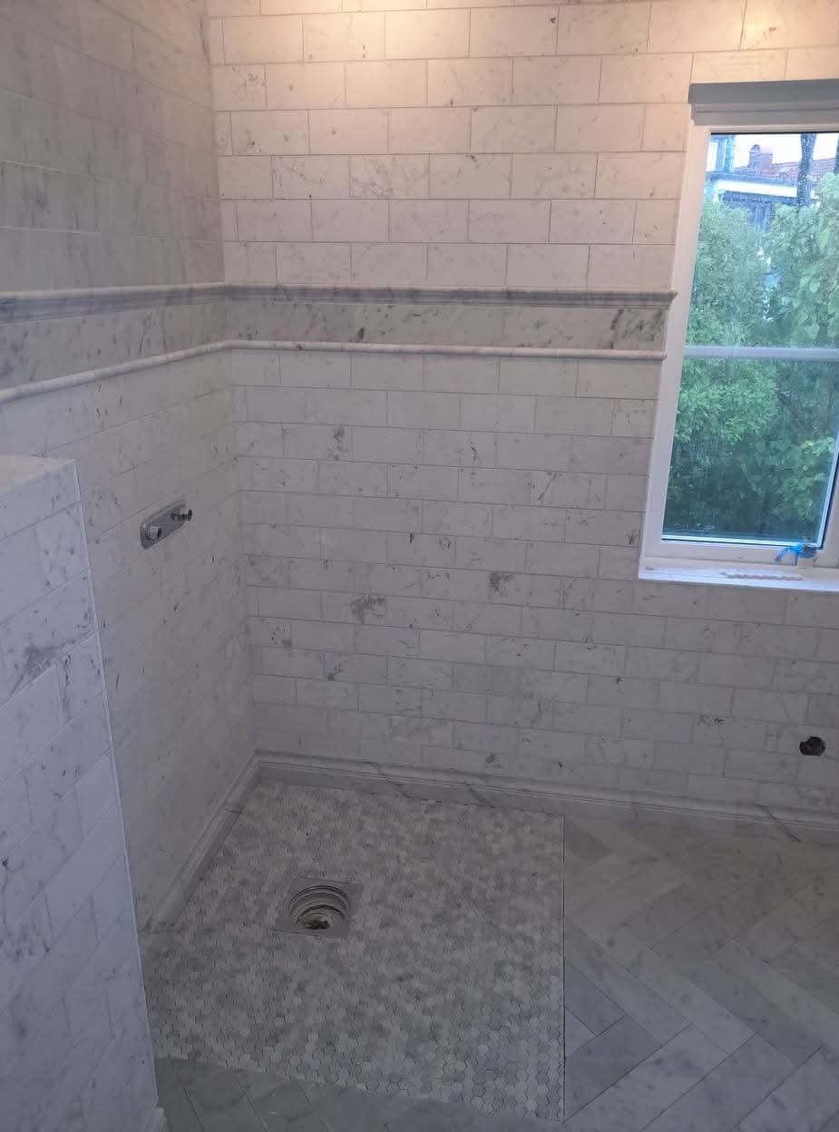 Wykończona łazienka z białymi marmurowymi płytkami ułożonymi w cegiełkę na ścianach i heksagonalną mozaiką pod prysznicem, widok na zewnątrz przez okno.