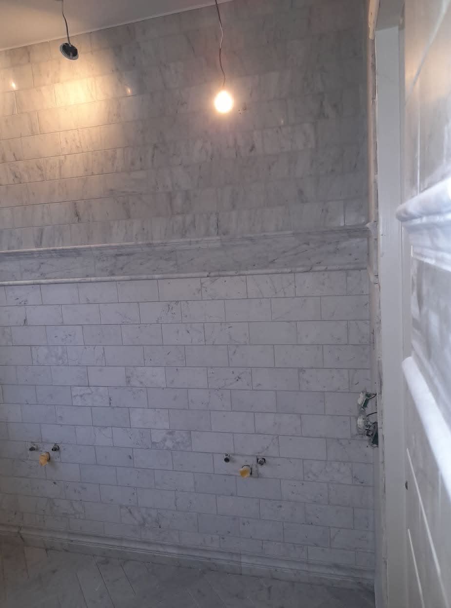 Ściana łazienki wyłożona marmurowymi płytkami w odcieniach szarości, z wystającymi rurami i przewodami elektrycznymi, oświetlona wiszącymi żarówkami, w trakcie prac wykończeniowych.