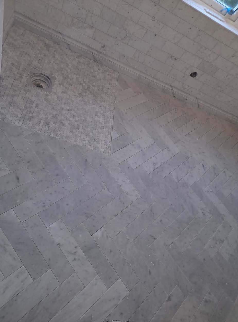 Ukończona podłoga łazienki wyłożona szarymi płytkami w układzie jodełki, z odpływem liniowym wykończonym mozaiką heksagonalną.