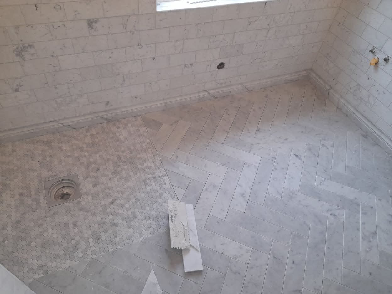 Ukończona łazienka z białymi marmurowymi płytkami ułożonymi w jodełkę na podłodze i prostokątnie na ścianach, widoczny odpływ i szpachelka z zaprawą.