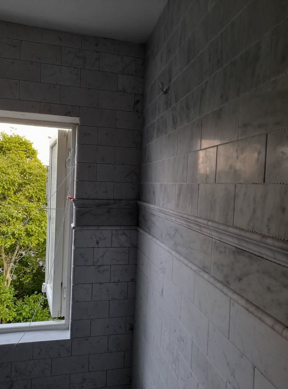 Ściana wyłożona marmurowymi płytkami z dekoracyjną listwą, widok na otwarte okno z zielenią na zewnątrz.