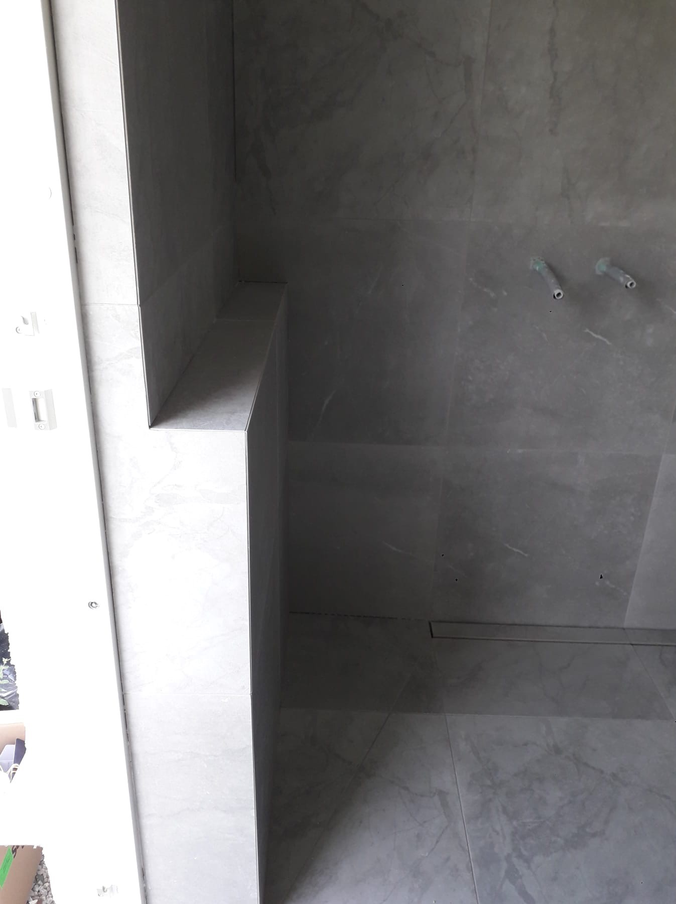 Szara, marmurkowa glazura ścienna w łazience, z widocznymi rurami instalacji wodnej i odpływem liniowym w podłodze, półka wyłożona płytkami.