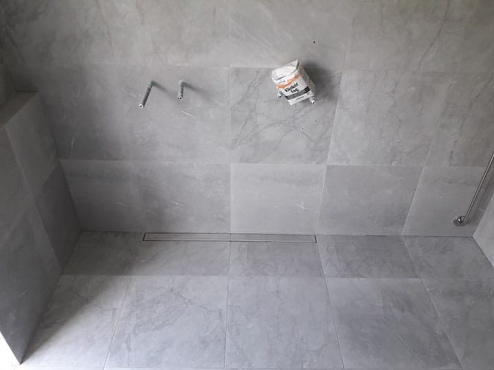 Łazienka w trakcie wykańczania: szare, marmurowe płytki, liniowy odpływ podłogowy, wystające rury instalacji wodnej, worek z klejem do płytek oparty o ścianę.