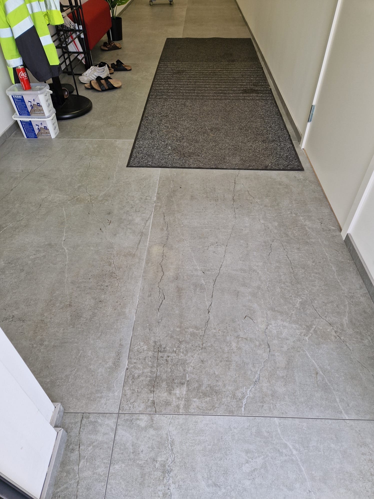 Szare płytki podłogowe imitujące kamień z ciemnoszarym dywanikiem przy drzwiach, widoczne buty, puszka Coca-Coli i odblaskowa kamizelka.