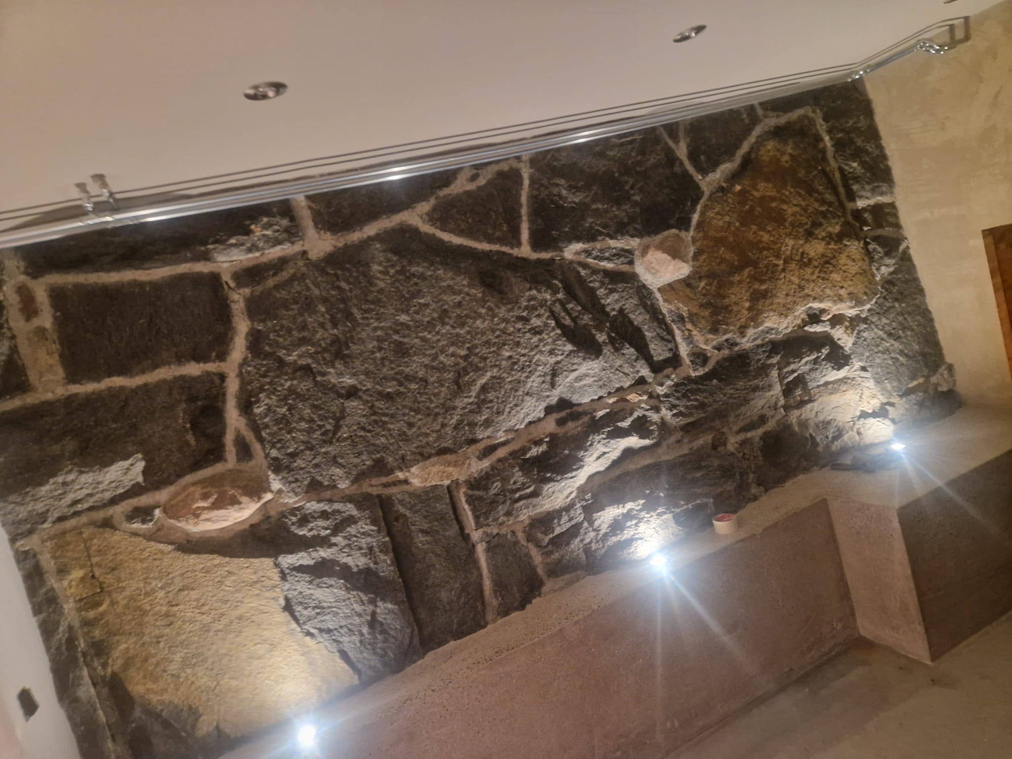 Ściana z nieregularnych kamieni o zróżnicowanej fakturze, oświetlona punktowymi lampami, z widoczną instalacją sufitową.