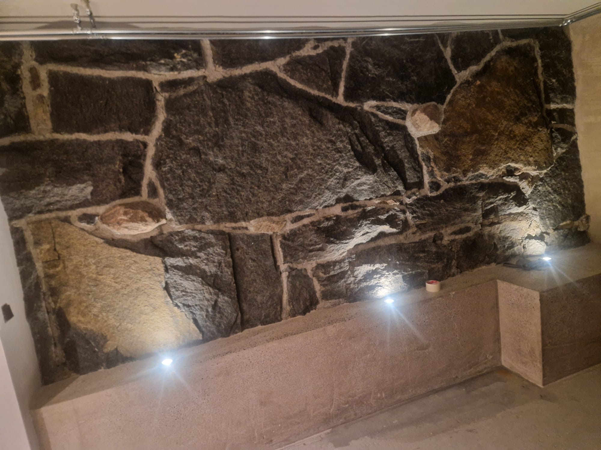 Dekoracyjna ściana z nieregularnych kamieni oświetlona punktowymi lampami LED w dolnej części, z widoczną instalacją pod sufitem.