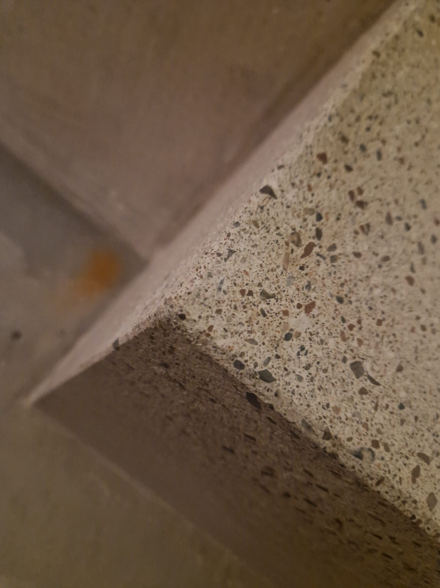 Fragment narożnika ściany wykończonej lastryko z widocznymi drobnymi kamyczkami, oświetlony z góry, z pomarańczowym przebarwieniem na sąsiedniej powierzchni.