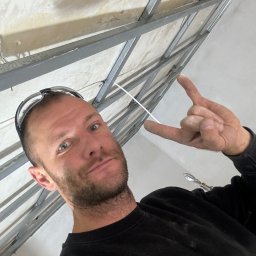 MATEUSZ SAMBORSKI SAMBUD - Mężczyzna w okularach ochronnych pokazuje znak rogów na tle metalowej konstrukcji sufitu podwieszanego z płyty karton gips, widoczne narzędzia.