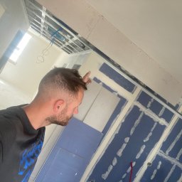 MATEUSZ SAMBORSKI SAMBUD - Mężczyzna wskazujący na sufit podwieszany z płyt karton gips, widoczne profile, instalacja elektryczna i niebieskie płyty gipsowe ze śladami szpachlowania.