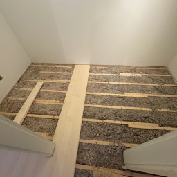 MATEUSZ SAMBORSKI SAMBUD - Ocieplenie podłogi wełną mineralną między legarami, częściowo zakryte deskami. Widok z góry na poddasze lub strych w trakcie prac wykończeniowych.