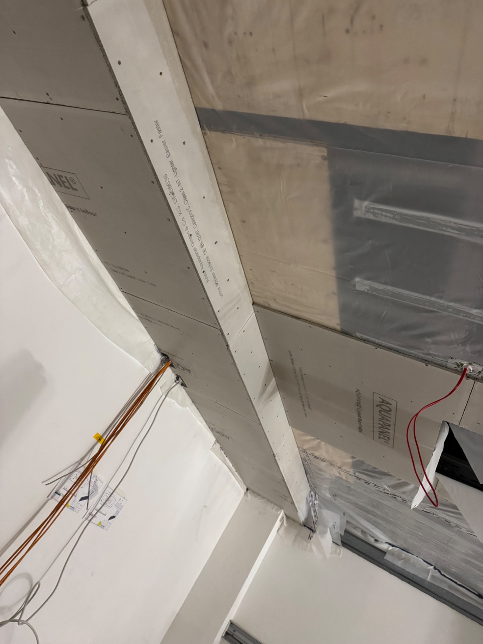 Montaż płyt Aquapanel na suficie, widoczne profile, folia ochronna, kable elektryczne i plany budowlane przy ścianie.