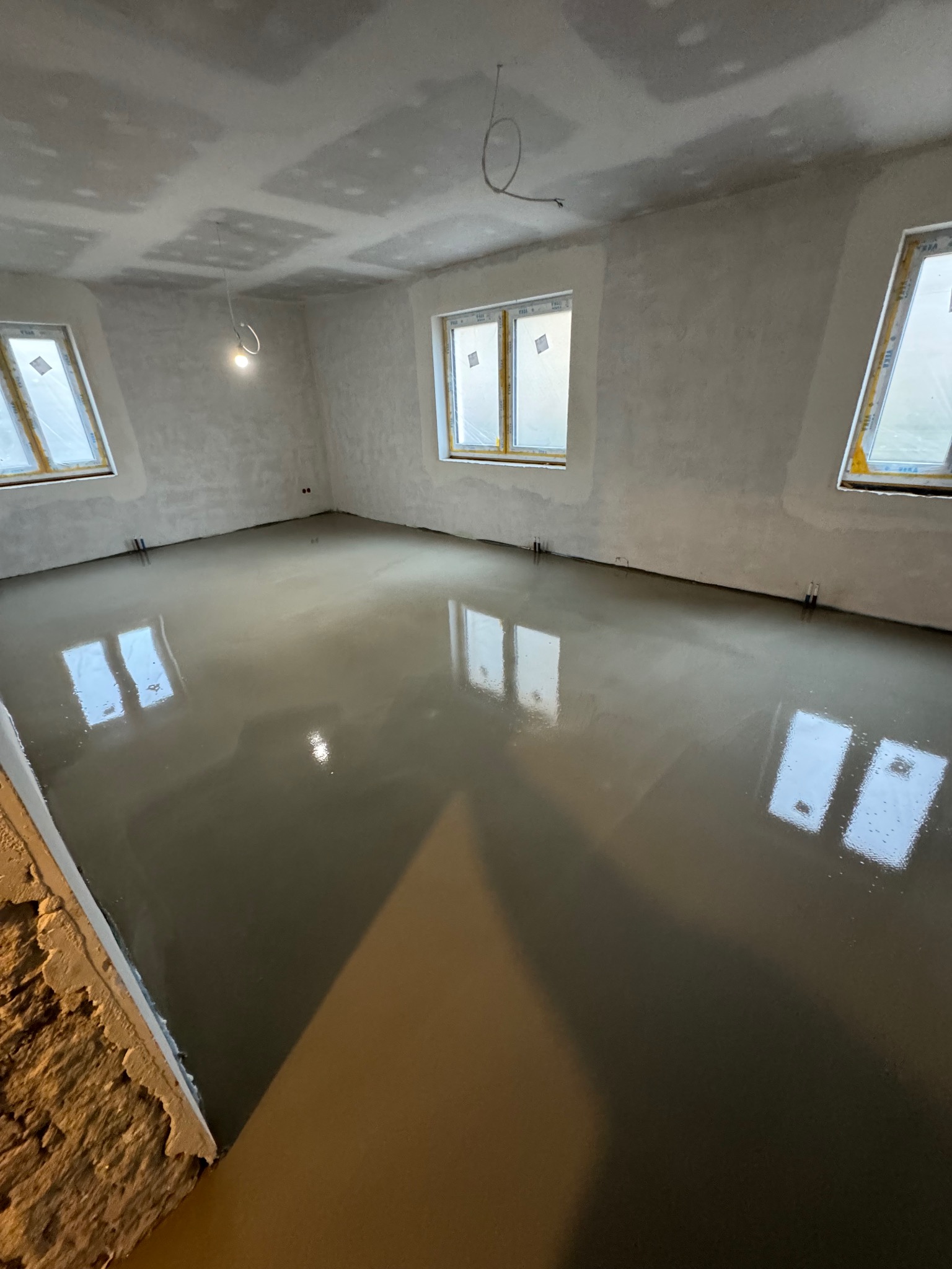 Wylewka samopoziomująca na podłodze w pomieszczeniu z oknami zabezpieczonymi folią, ściany przygotowane pod gładzie, widoczne kable oświetleniowe.