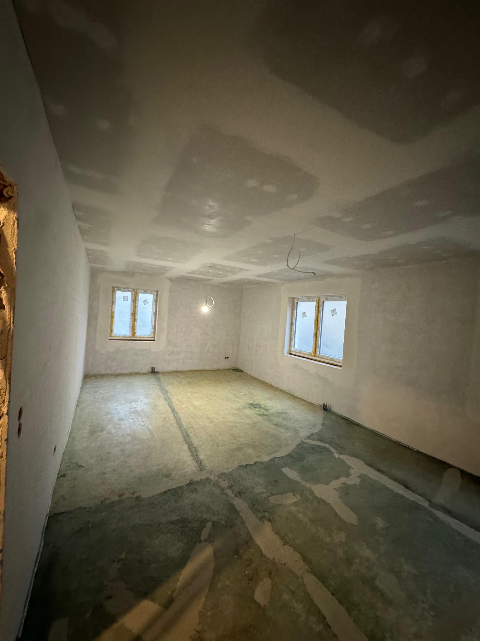 Pokój w trakcie prac wykończeniowych: ściany i sufit po wstępnym szpachlowaniu, okna zabezpieczone folią, betonowa wylewka na podłodze z widocznymi śladami wilgoci.