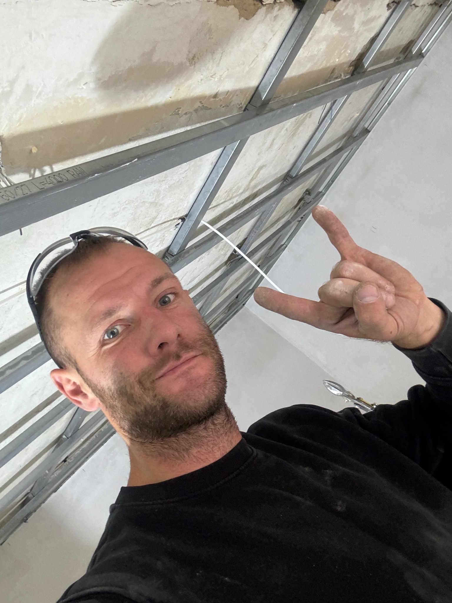 Mężczyzna w okularach ochronnych pokazuje znak rogów na tle metalowej konstrukcji sufitu podwieszanego z płyty karton gips, widoczne narzędzia.