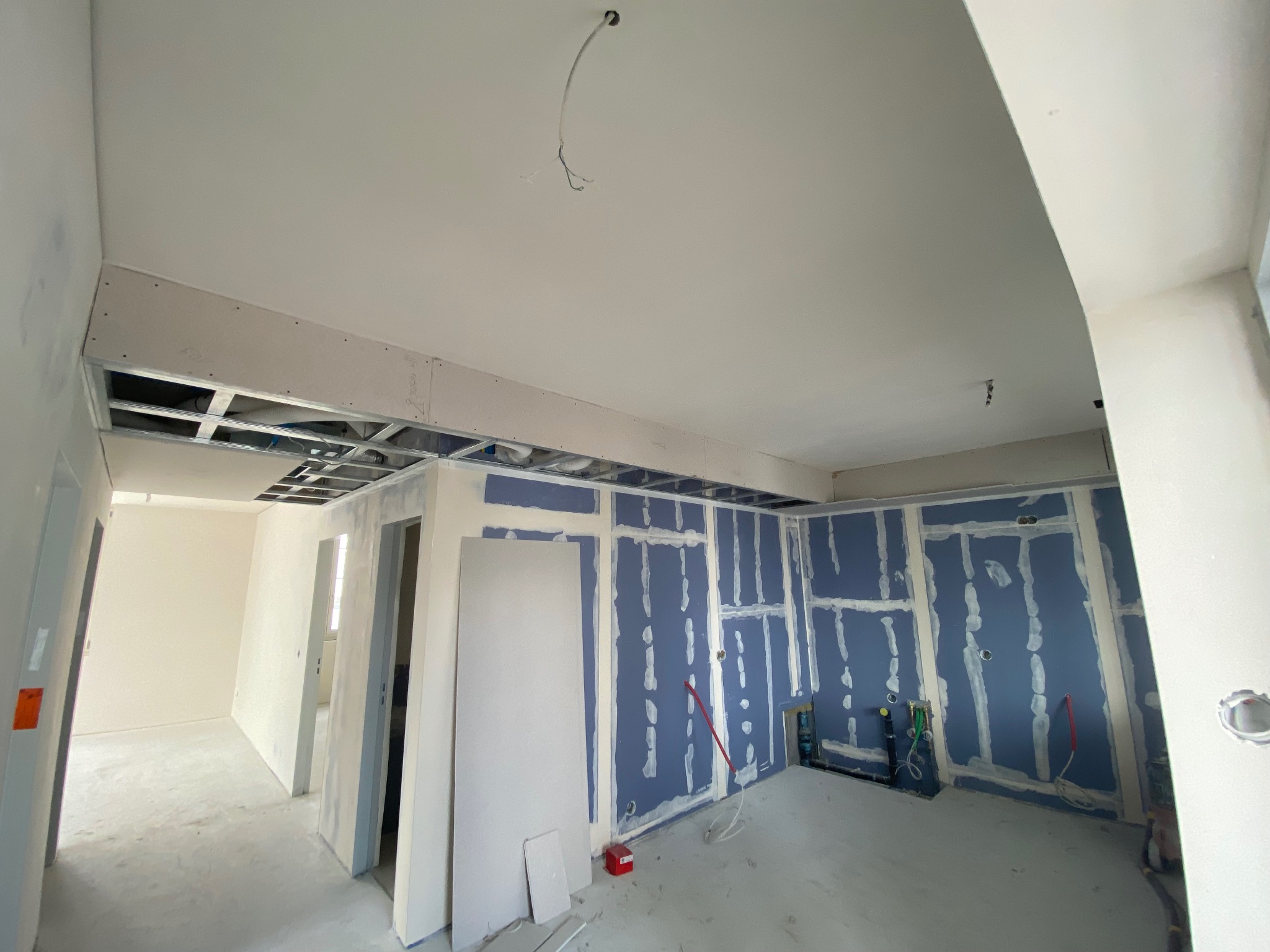 Wnętrze w trakcie prac, widoczne metalowe profile sufitowe, niebieska ściana z nałożoną siatką i gipsem, przygotowana pod dalsze prace wykończeniowe, oraz płyty karton-gips oparte o ścianę.