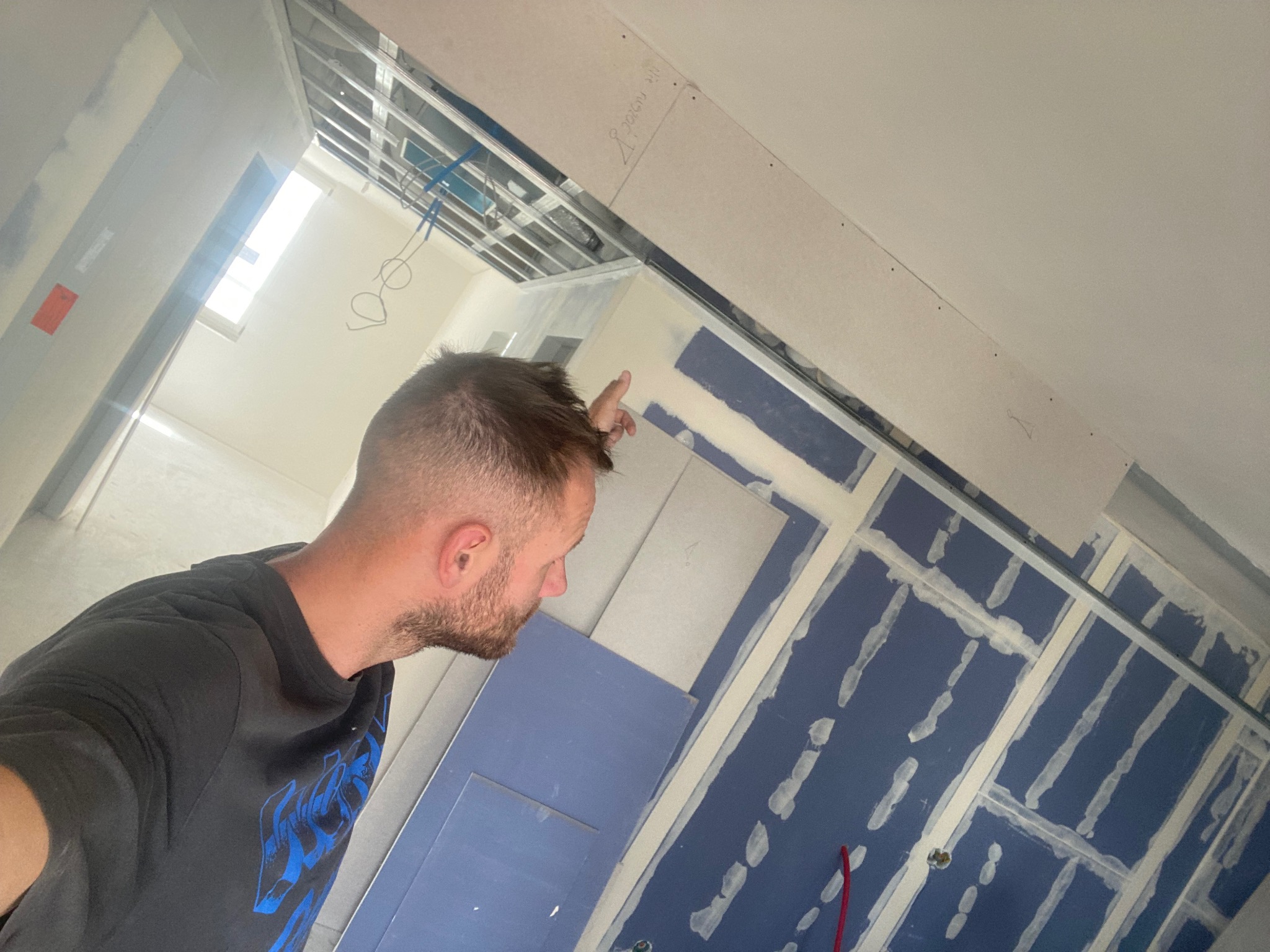 Mężczyzna wskazujący na sufit podwieszany z płyt karton gips, widoczne profile, instalacja elektryczna i niebieskie płyty gipsowe ze śladami szpachlowania.