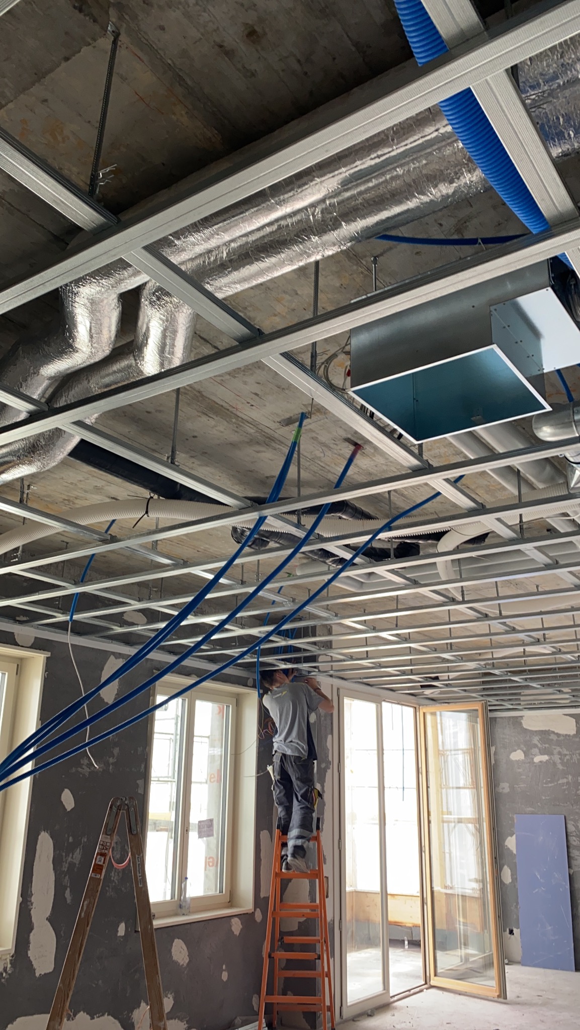 Montaż sufitu podwieszanego z widoczną konstrukcją metalową, izolacją termiczną, niebieskimi rurami wentylacyjnymi i pracownikiem na drabinie w surowym wnętrzu z oknami.