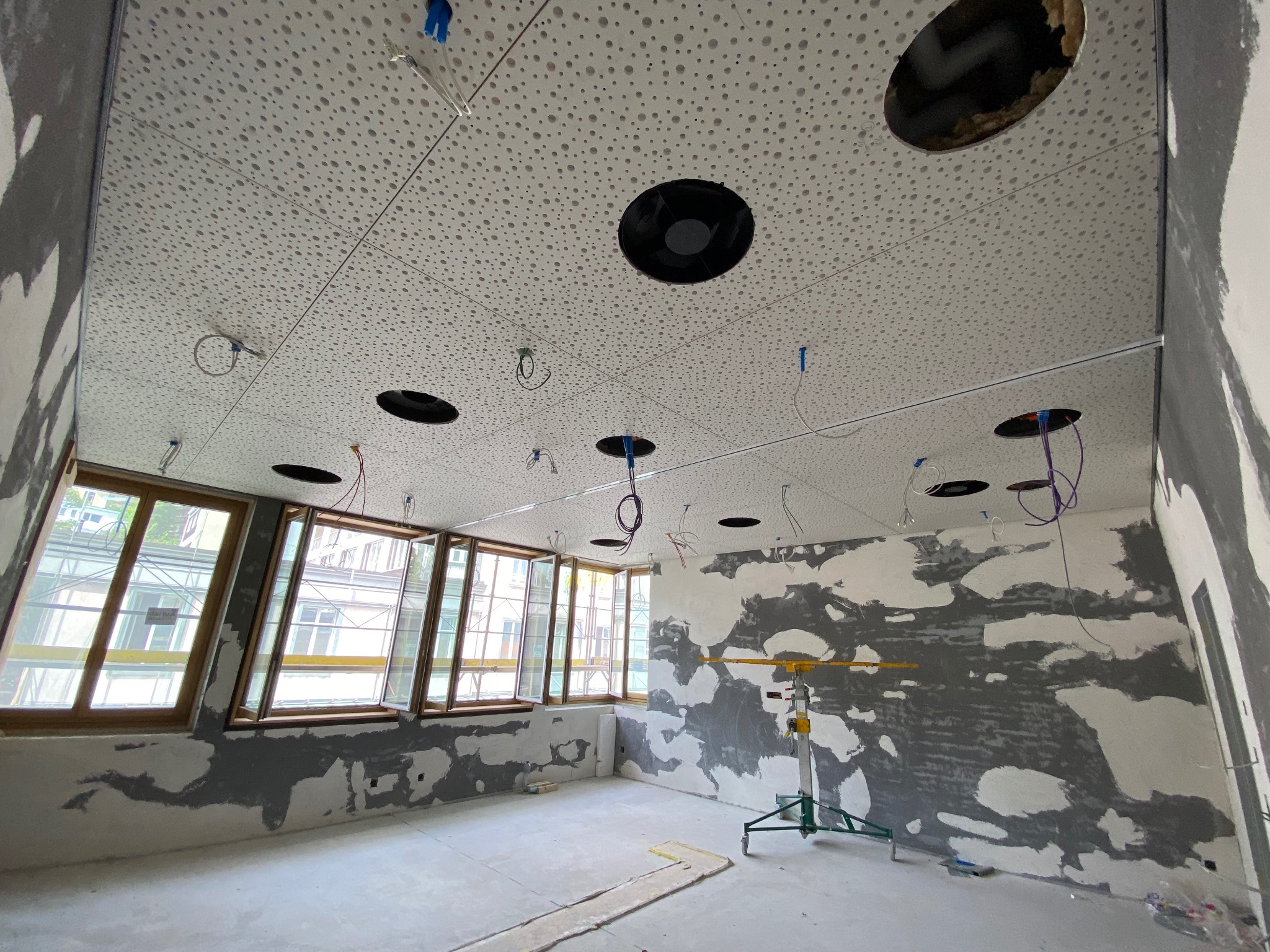 Wnętrze w trakcie prac wykończeniowych: sufit podwieszany z otworami na oświetlenie, ściany w trakcie szpachlowania, widoczne okna i podnośnik teleskopowy.