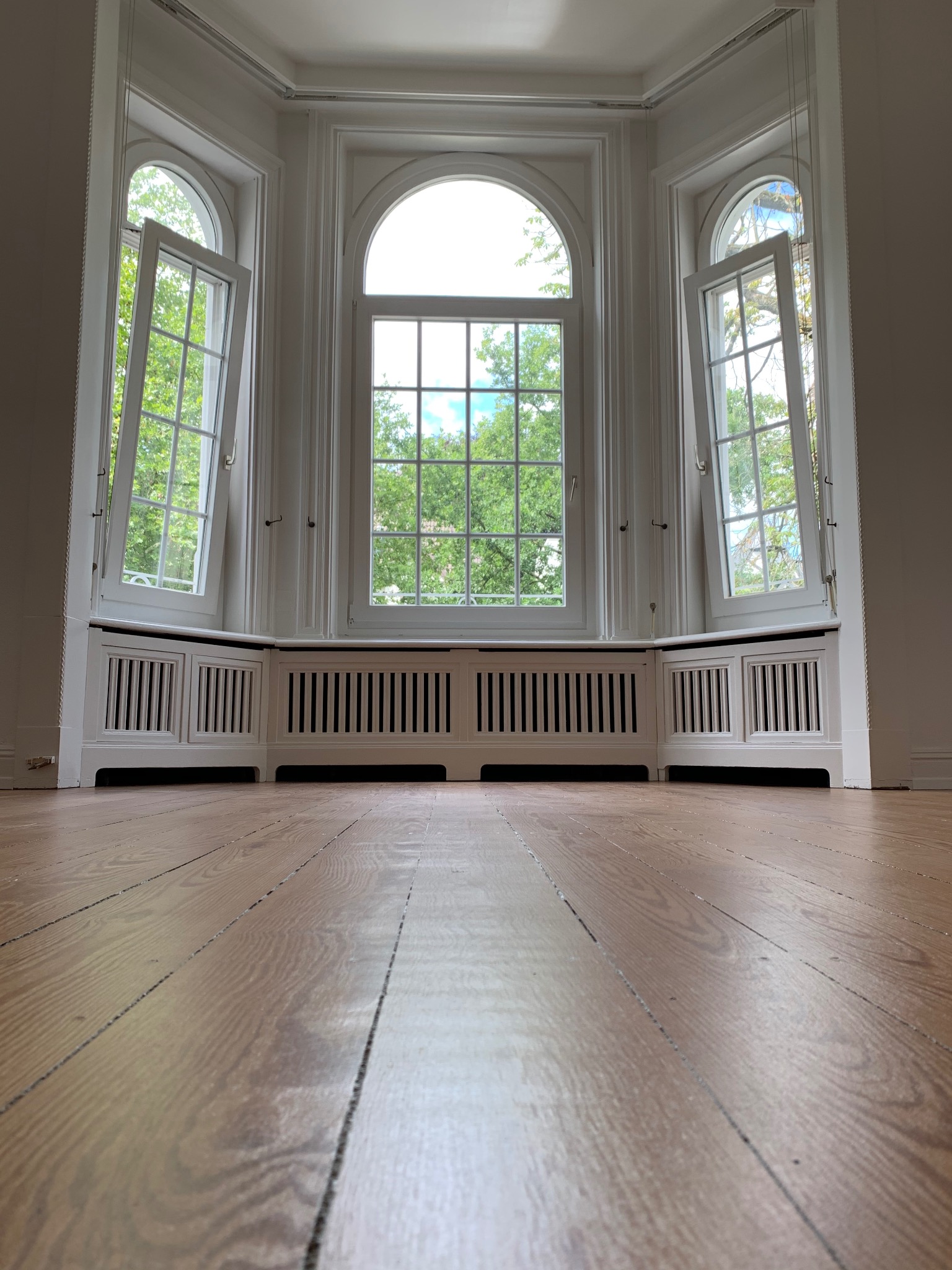 Pusty pokój z odnowioną podłogą z desek, trzema wysokimi oknami z białymi ramami, z których dwa są otwarte, oraz białymi grzejnikami pod oknami.