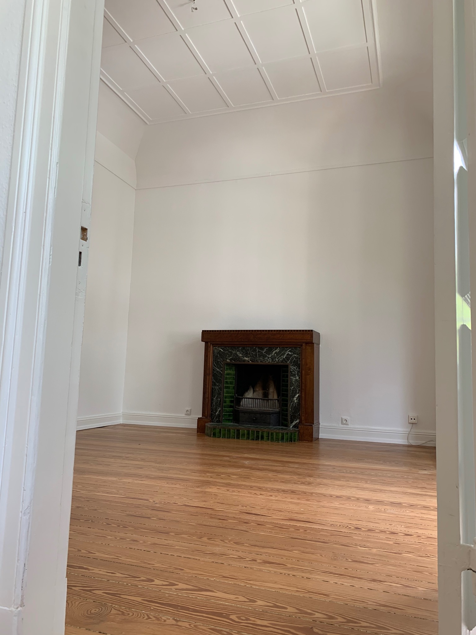 Pusty pokój z odnowioną podłogą z desek i ozdobnym kominkiem z zielonymi akcentami, widok zza białych drzwi.