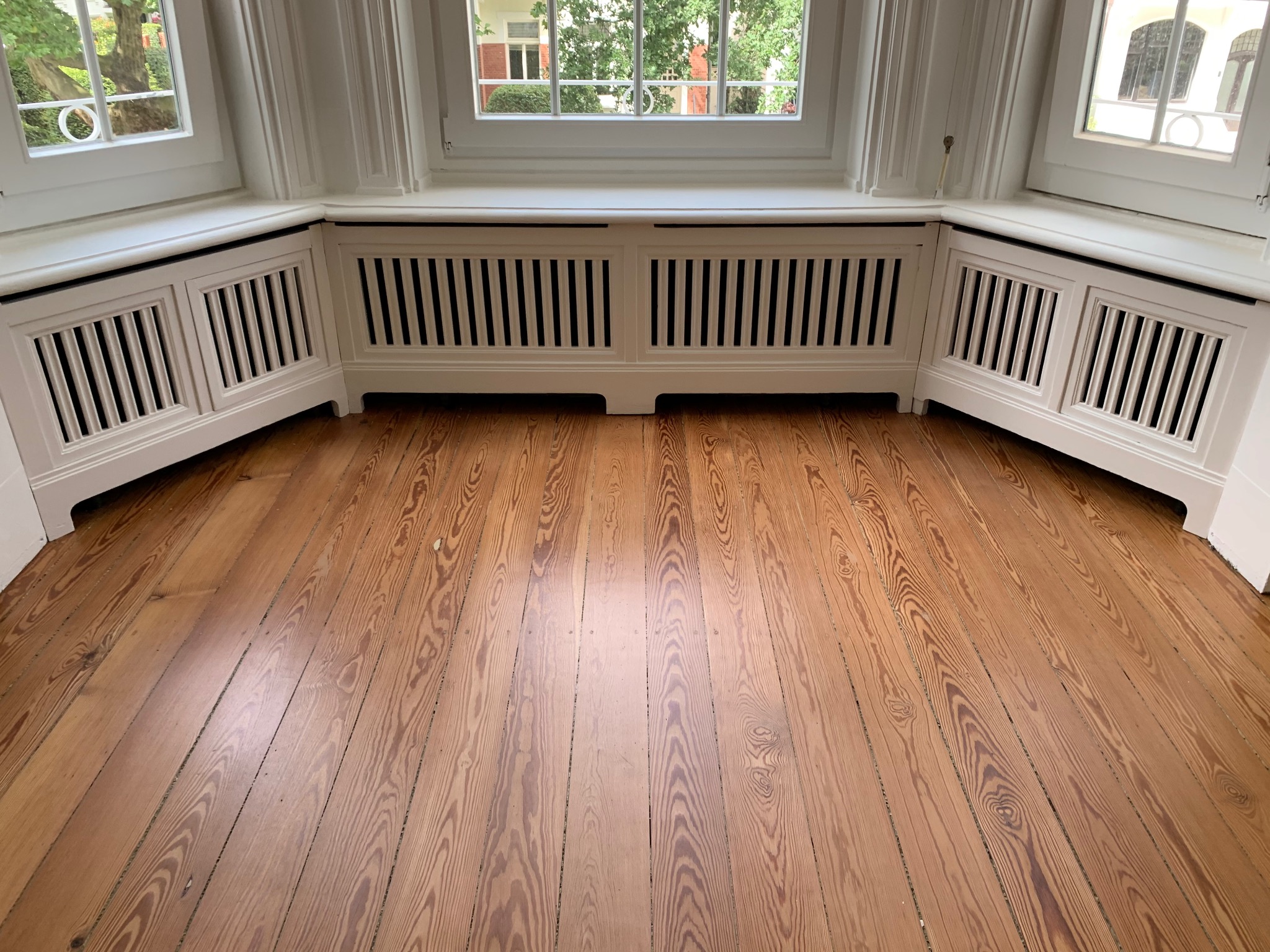 Jasne, lakierowane deski podłogowe z widocznym usłojeniem, białe obudowy grzejników pod oknami, wnętrze domu z widokiem na zieleń za oknem.