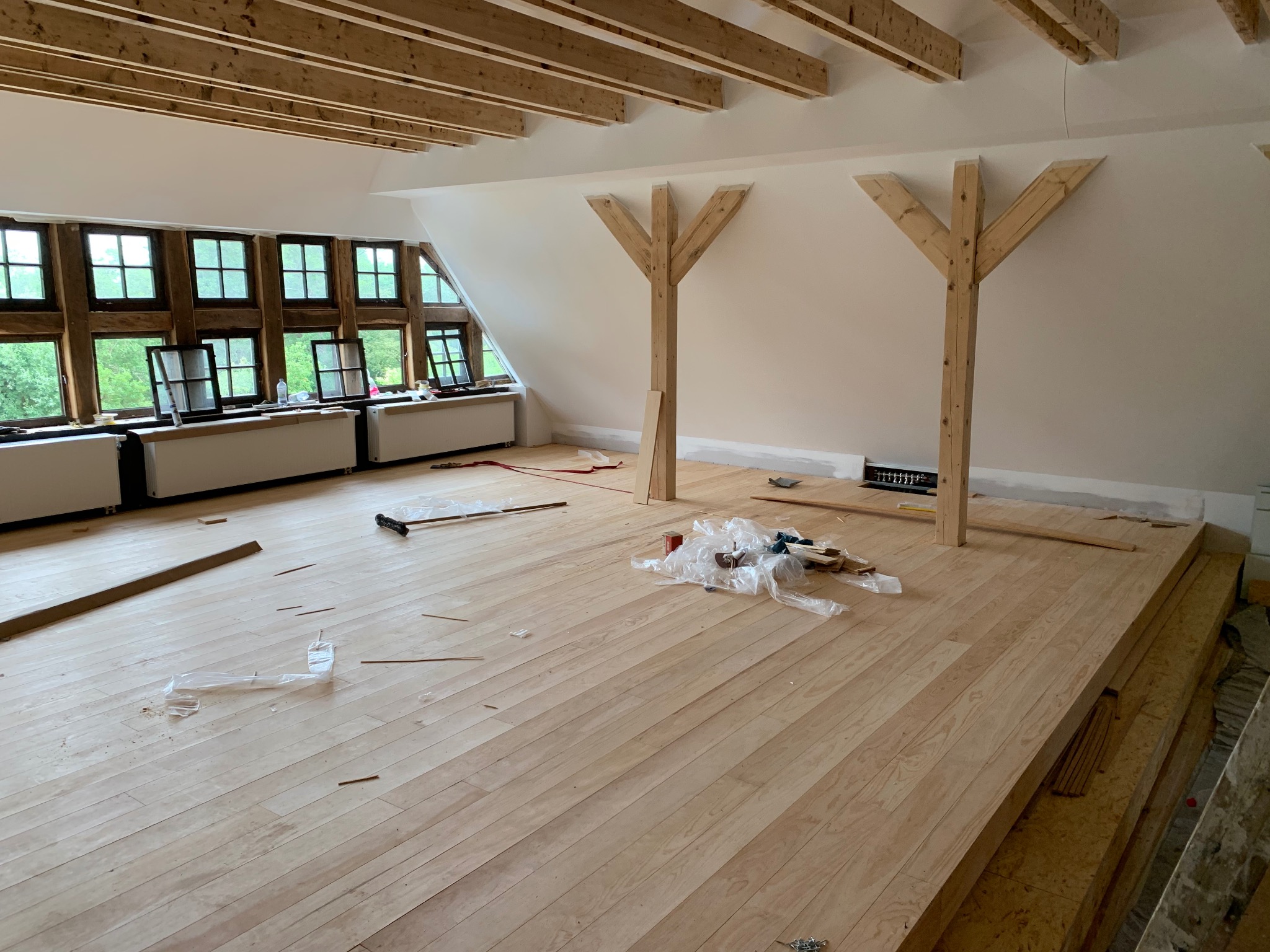 Poddasze w trakcie prac wykończeniowych: jasna drewniana podłoga, odsłonięte drewniane krokwie sufitowe, okna w drewnianych ramach, przygotowane drewniane podpory konstrukcyjne.