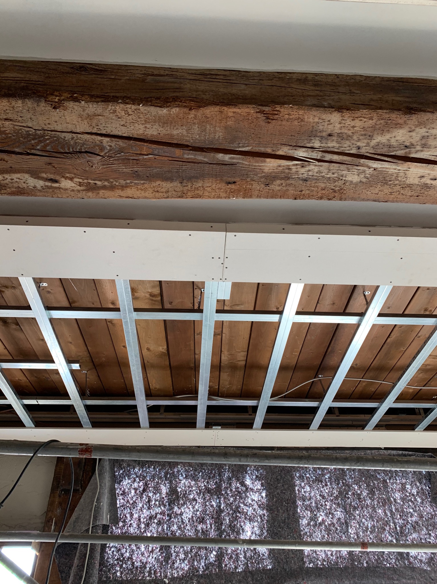 Montaż sufitu podwieszanego z płyt karton-gips na drewnianej konstrukcji. Widoczne metalowe profile i izolacja. Prace wykończeniowe w trakcie realizacji.