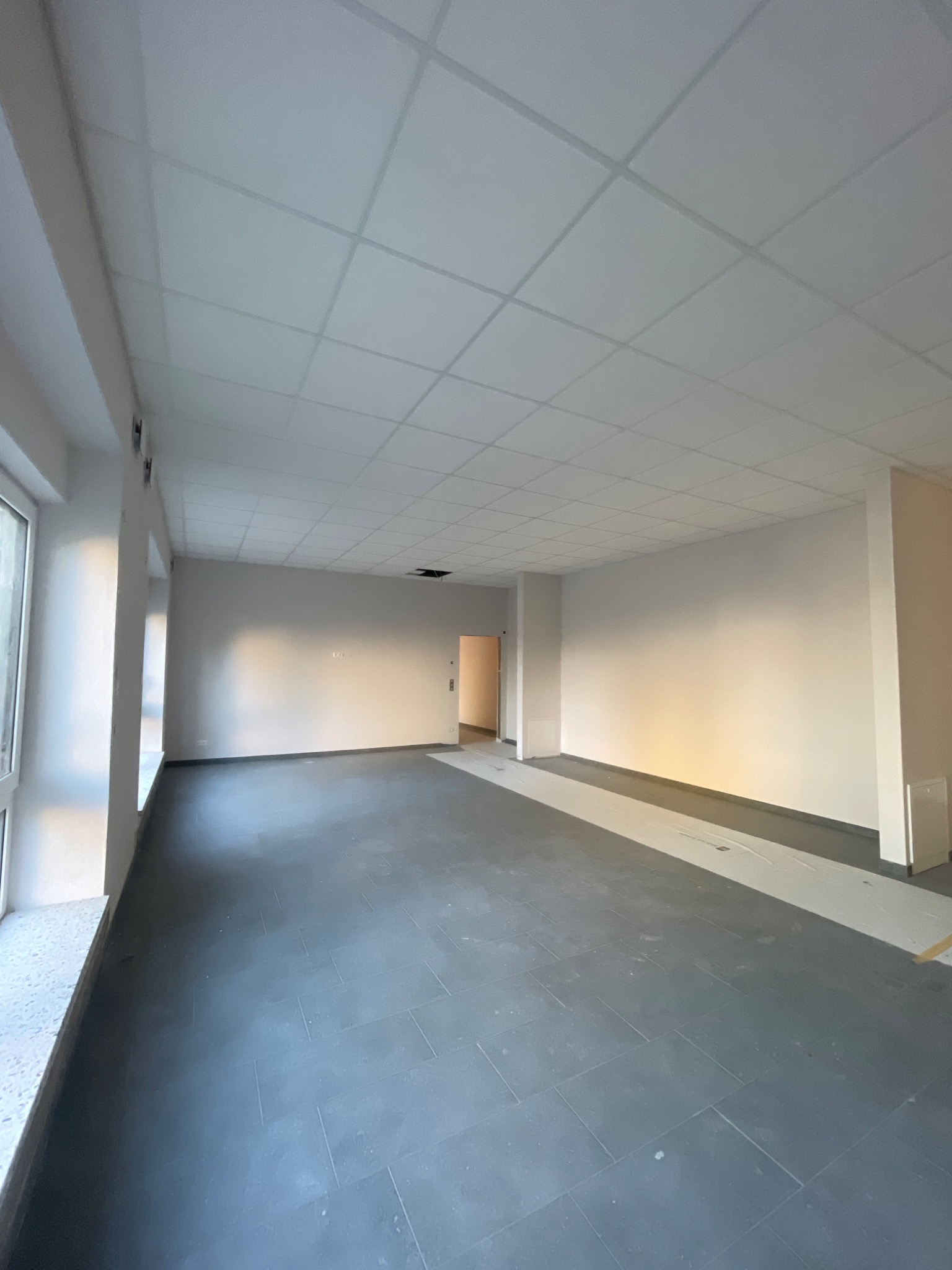 Wnętrze wykończonego biura z szarą podłogą, białymi ścianami i podwieszanym sufitem kasetonowym. Widoczne okna i drzwi w oddali.