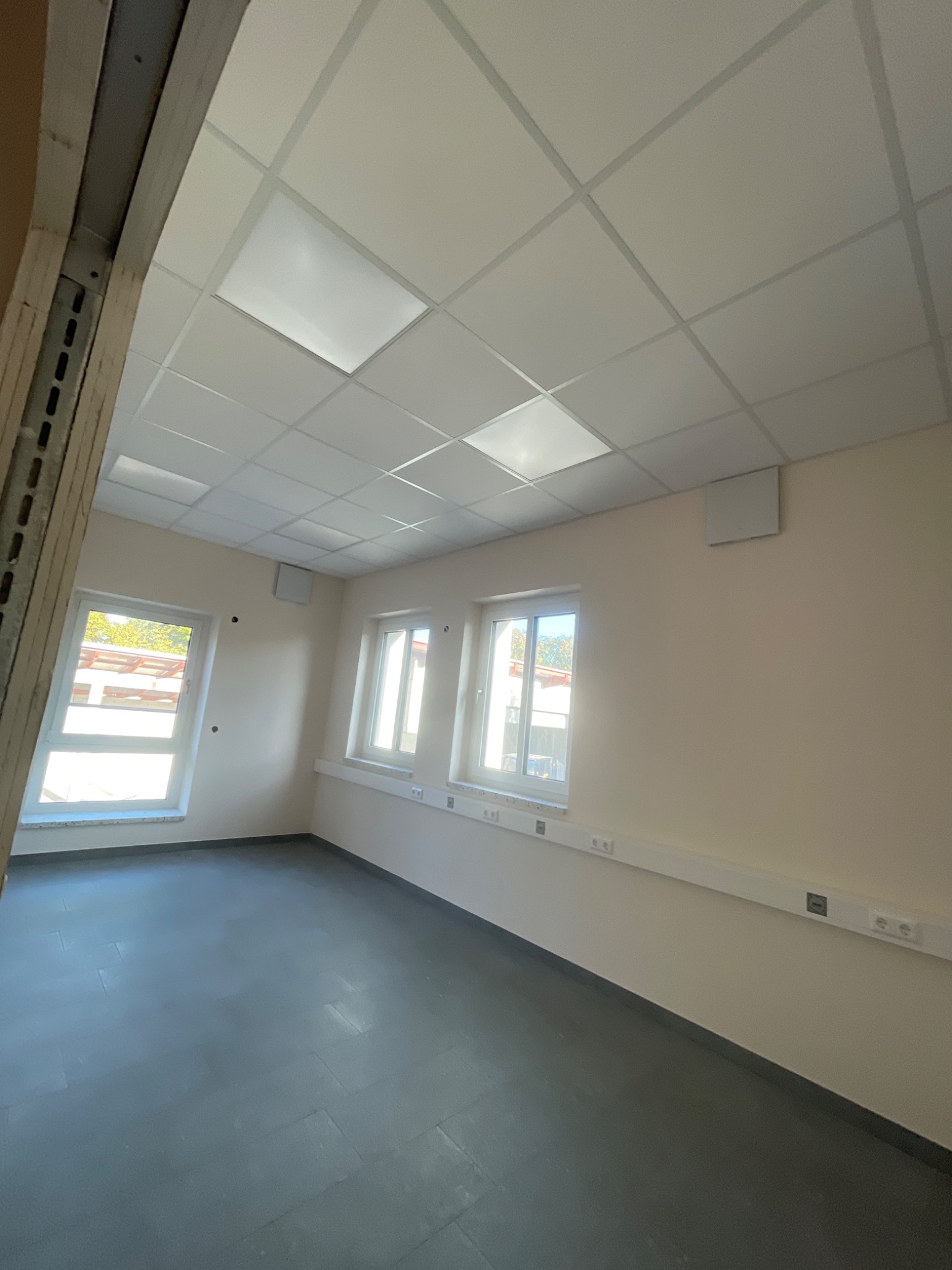 Wnętrze jasnego pokoju po remoncie z oknami, panelami podłogowymi i sufitem podwieszanym. Widoczne gniazdka elektryczne w listwie przypodłogowej.