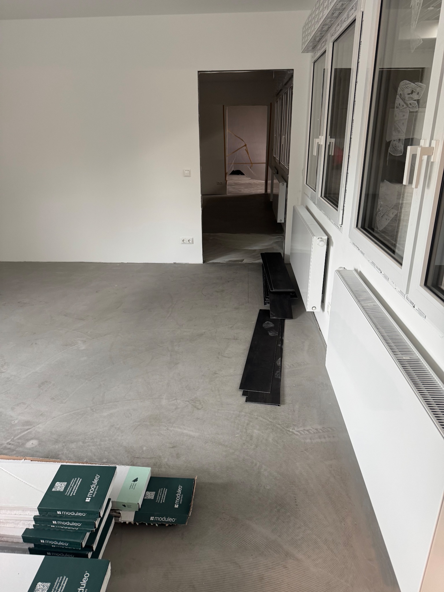 Przygotowanie do montażu paneli podłogowych: paczki paneli Moduleo, ułożone próbki, w tle wylewka, grzejniki i okna. Widok na wnętrze remontowanego pomieszczenia.