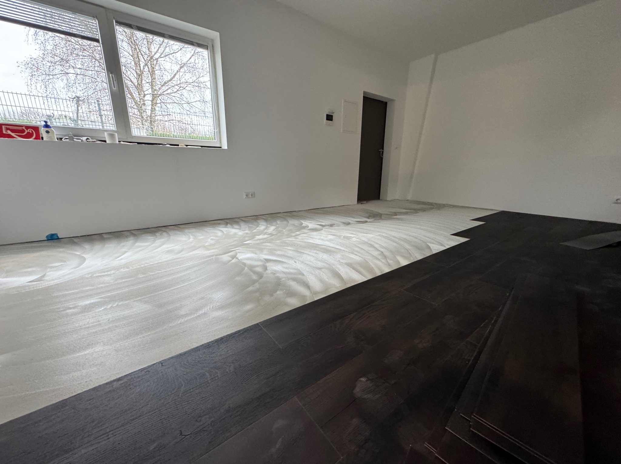 Układanie ciemnych paneli podłogowych w jasnym pokoju. Część podłogi pokryta klejem. Widok na okno i ścianę z drzwiami. Etap prac wykończeniowych.