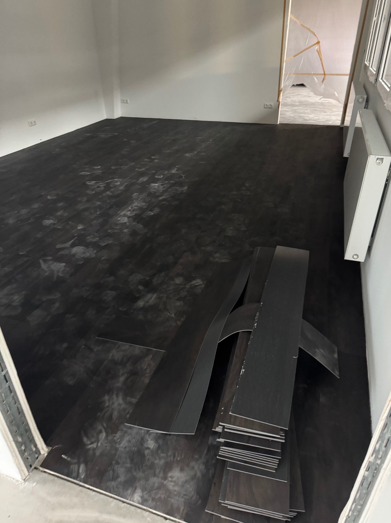 Ciemna podłoga z paneli winylowych w trakcie montażu. Widoczne ślady po butach i stos paneli przy ścianie. Jasne ściany i grzejnik. Ujęcie z niskiego kąta.