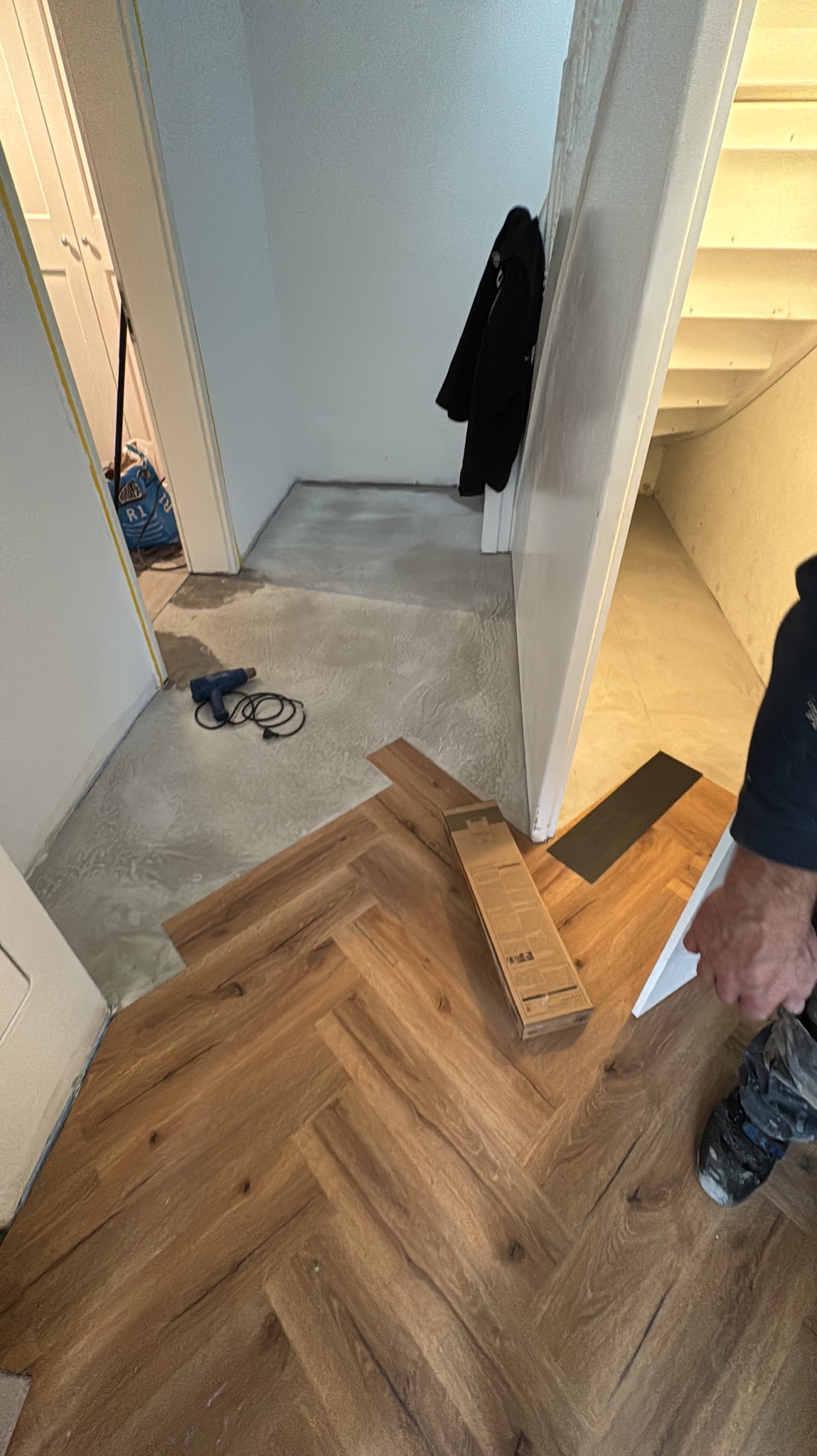 Układanie paneli w jodełkę w korytarzu, widoczny pistolet na gorące powietrze i paczka paneli. Częściowo ułożona podłoga, w tle schody i wieszak na ubrania.
