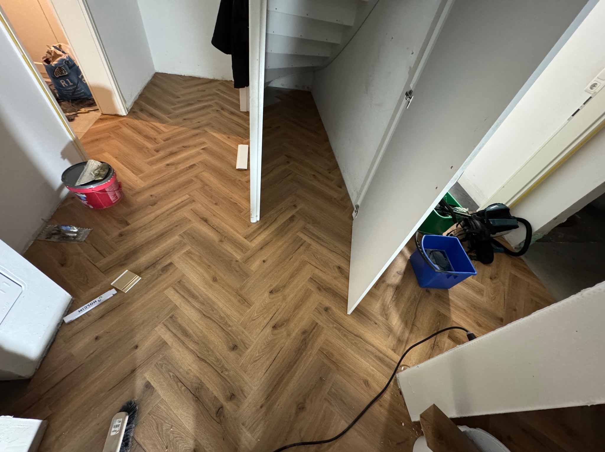 Ukończona podłoga z paneli ułożonych w jodełkę, widok z góry, z elementami narzędzi i materiałów budowlanych w tle. Jasne wnętrze, częściowo widoczne schody.